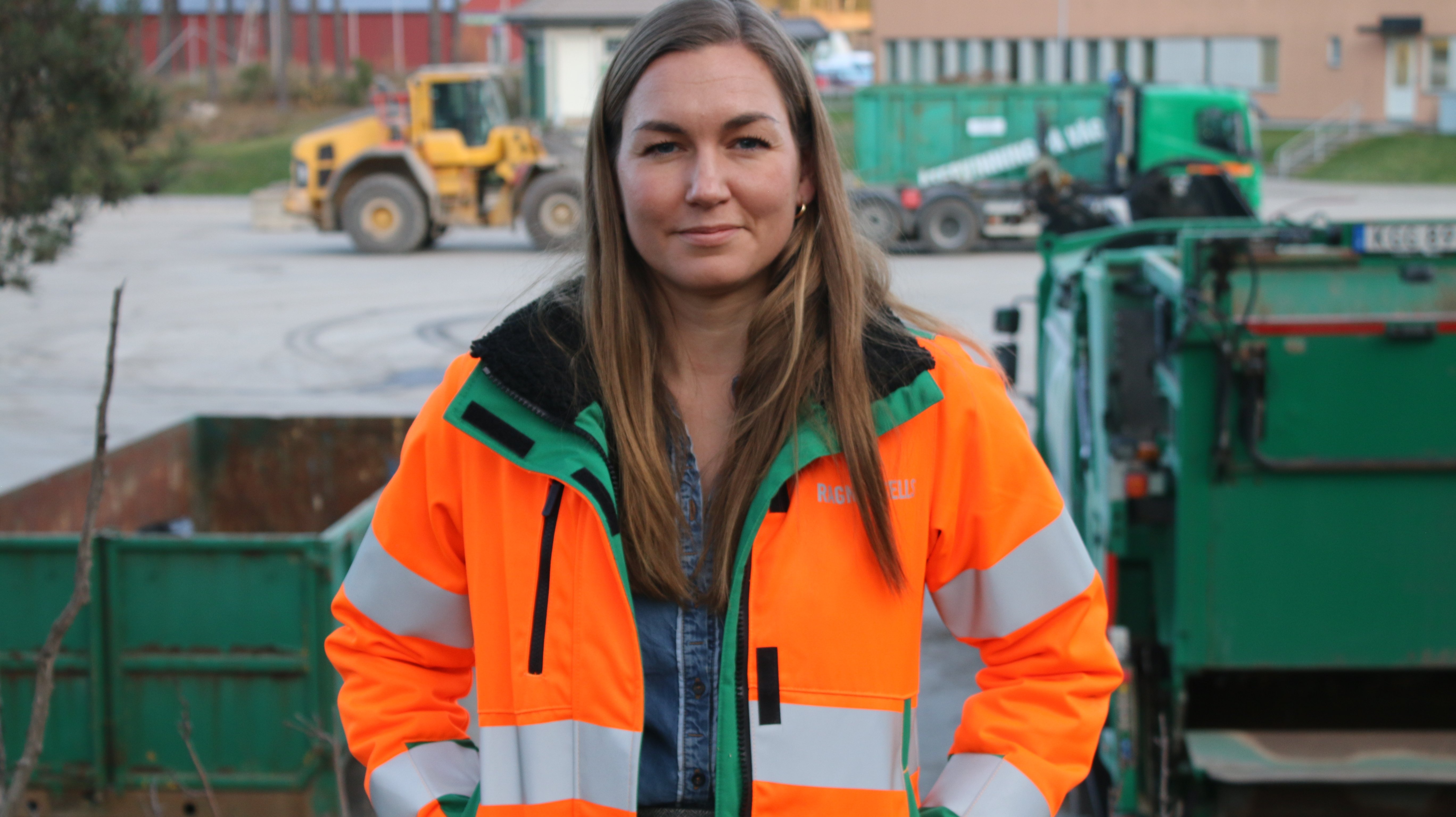 Julia står med orange jacka, i bakgrunden finns fordon