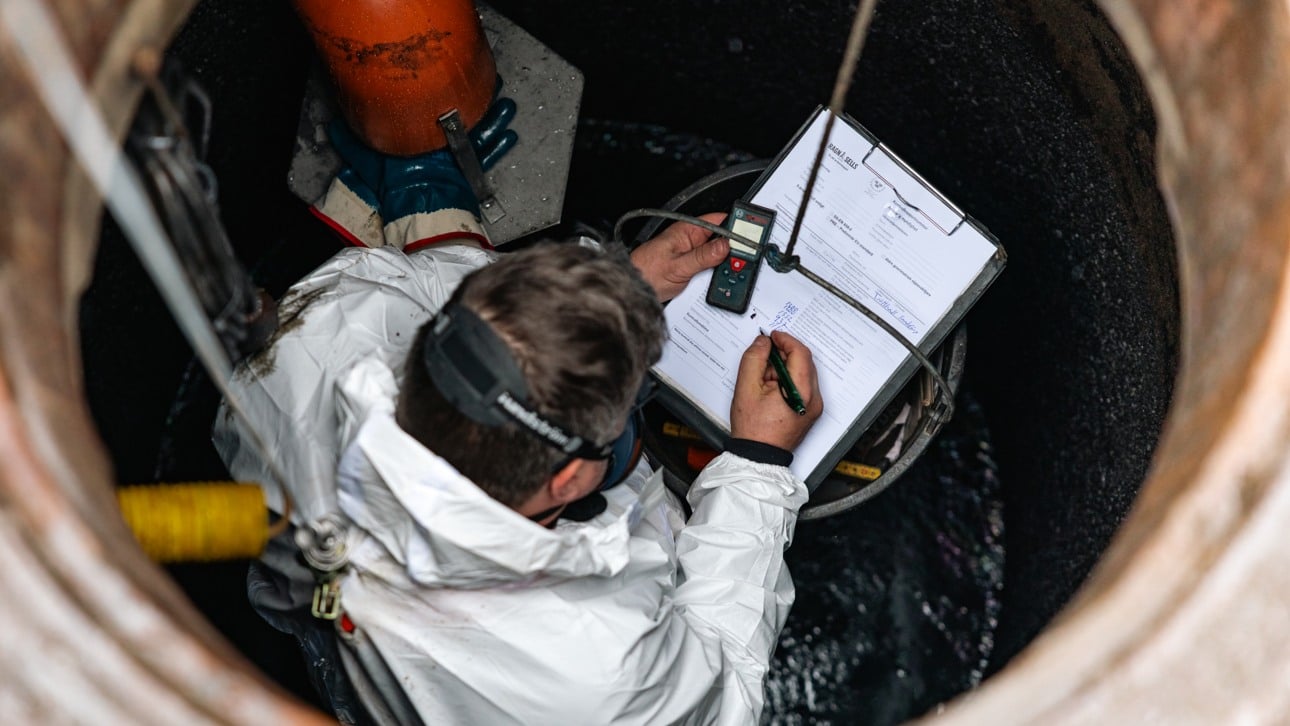 Man från Ragn-Sells i vit skyddsdräkt och munskydd nere i en Oljeavskiljare och fyller i protokoll